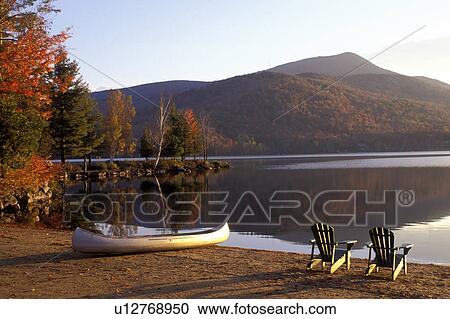 Stock Image -  Adirondack chairs, lake, canoe, Adirondacks, New York, Blue Mountain Lake, NY, A canoe and two Adirondack chairs on a quiet beach on the calm waters of Blue Mountain Lake in the autumn in the Adirondacks.. Fotosearch