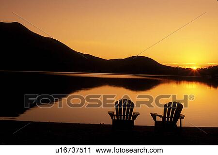 Stock Image -  Adirondack chairs, sunrise, lake, Adirondacks, New York, Blue Mountain Lake, NY, A silhouette of two Adirondack chairs on the beach at sunrise on Blue Mountain Lake in the Adirondacks.. Fotosearch