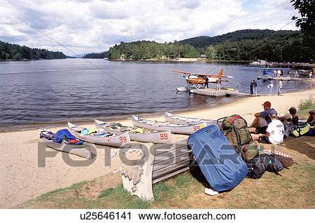 Stock Image -  Adirondacks, New York, NY, Long Lake, Canoes, people resting & seaplane along the beach at Long Lake in the Adirondack Mountains.. Fotosearch