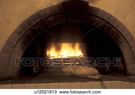 Stock Image -  bakery, bread, brick oven, fire, flame, wood stove, Wood burning in a brick oven with a loaf of freshly baked bread displayed in front of the oven at Gerard's Bakery in Westford in the state of Vermont. . Fotosearch
