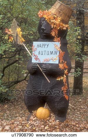 Stock Image -  bear, autumn, fall decoration, decorations, Large wooden bear sculpture displays a Happy Autumn sign dressed with a basket of colorful maple leaves on his head and rake in hand for fall display in Arlington in Bennington County in the state of Vermont.. Fotosearch