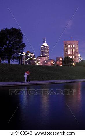 Stock Image -  canal, Indianapolis, skyline, park, IN, Indiana, A couple walks along Historic Central Canal in the evening with the view of the downtown skyline in the distance in Indianapolis.. Fotosearch