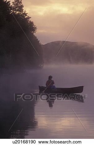 Stock Image -  canoeing, canoe, sunrise, sunset, Vermont, VT, Woman paddling a canoe on Mollys Falls Pond at sunrise in the fog.. Fotosearch
