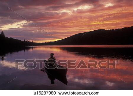 Stock Image -  canoeing, canoe, sunset, sunrise, Vermont, VT, Woman paddling a canoe on Kettle Pond at sunset in Groton State Forest.. Fotosearch