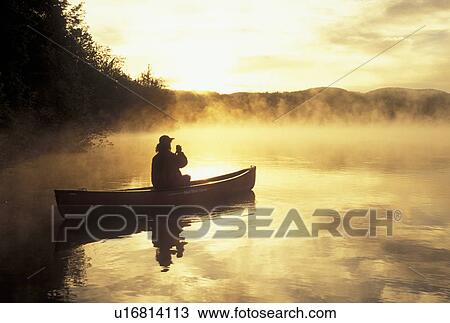 Stock Image -  canoeing, canoe, sunset, sunrise, Vermont, VT, Woman paddling a canoe on Mollys Falls Pond at sunrise in the mist.. Fotosearch
