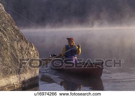 Stock Photograph -  canoeing, canoe, Vermont, VT, Woman paddles a canoe on Kettle Pond through boulders in the mist in Groton State Forest.. Fotosearch