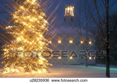 Stock Photograph -  church, Christmas tree, chapel, decorations, holiday, outdoor, natural, winter, town, snow, Vermont, A large evergreen tree with bright white starlights and candles in the windows of the Old Round Church (a 16-sided polygon) National Historical Landmark . Fotosearch