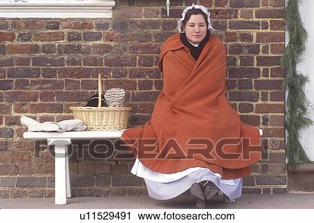 Stock Image -  Colonial Williamsburg, Virginia, VA, Williamsburg, A woman interpreter dressed in a colonial costume sits outside the Courthouse in Colonial Williamsburg.. Fotosearch