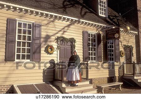 Stock Image -  Colonial Williamsburg, Virginia, VA, Williamsburg, Interpreter wearing colonial costume stands outside the village store in Colonial Williamsburg.. Fotosearch