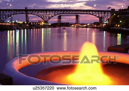 Stock Image -  fountain, bridge, Cleveland, OH, Ohio, Flats District, Fountain at Settlers Landing Park along the Cuyahoga River in the Flats District of downtown Cleveland illuminated at night. Main Avenue Bridge crossing the Cuyahoga River in the background.. Fotosearch