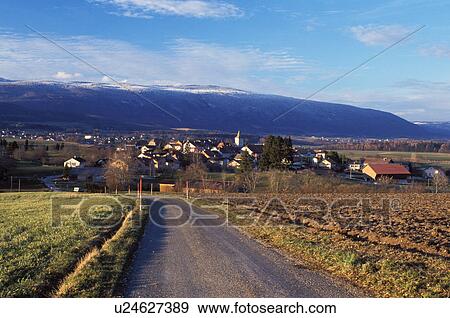 Stock Photo -  Switzerland, Vaud, Essertines-s-Rolle, Jura Mountains, Europe, A country road leads down to the village of Essertines/Rolle with a view of the snow covered Jura Mountains in the background in winter.. Fotosearch