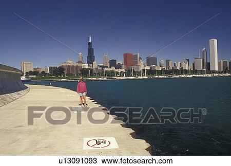 Stock Image -  Chicago, IL, lakeshore, Illinois, Lake Michigan, skyline, Scenic view of Chicago Harbor and the downtown skyline from Grant Park in Chicago. Woman walking along the lakeshore recreational path.. Fotosearch