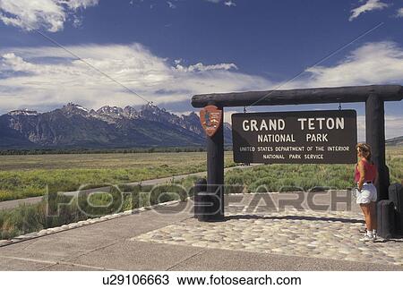 Stock Image -  Grand Teton National Park, WY, Wyoming, Woman reading sign at the entrance to Grand Teton Nat'l Park with a scenic view of the Grand Teton Mountains in Wyoming.. Fotosearch