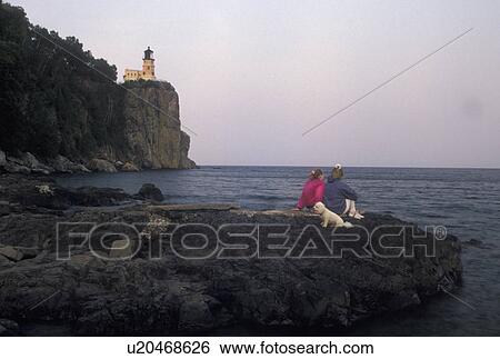 Stock Photograph -  lighthouse, Lake Superior, North Shore Drive, Two Harbors, MN, Minnesota, People with their pet dog sitting on a large rock along the north shore looking at Split Rock Lighthouse situated on a high cliff on Lake Superior at Split Rock Lighthouse State Pa. Fotosearch