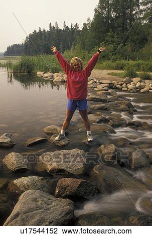 Stock Image -  Mississippi River, Itasca State Park, MN, Minnesota, Park Rapids, Lake Itasca, A woman stands with hands in the air after crossing the narrow headwaters of the Mississippi River on large rocks in Itasca State Park.. Fotosearch