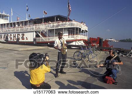 Stock Image -  street artist, New Orleans, Mississippi River, Riverfront Park, LA, Louisiana, Men record a street musician playing his instrument on Waldenberg Riverfront Park in front of the Steamboat Natchez along the Mississippi River in New Orleans. The Natchez is . Fotosearch