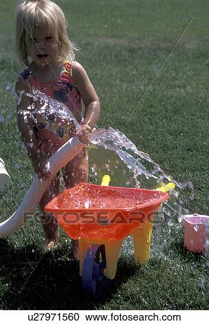 Stock Image -  toddler, girl, MN, Minnesota, Alexandria, summer camp, Toddler playing with large hose filling a small wheel barrow with water on the lawn at Maple Lake in Alexandria in the summer. . Fotosearch