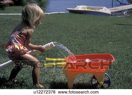 Stock Photo -  toddler, girl, MN, Minnesota, Alexandria, summer camp, Toddler playing with large hose filling a small wheel barrow with water on the lawn at Maple Lake in Alexandria in the summer. . Fotosearch