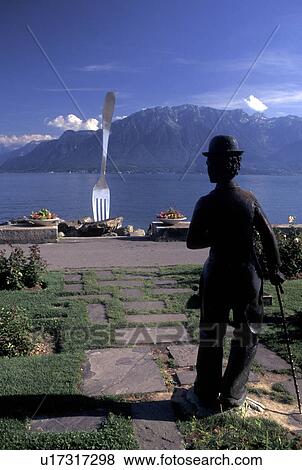 Stock Photo -  Charlie Chaplin, Vevey, Switzerland, Lake Geneva, Alps, Vaud, Charlie Chaplin statue and sculpture of a huge fork along the lakefront park along Lac Leman in Vevey in the Canton of Vaud. Scenic view of the Alps across Lake Geneva.. Fotosearch