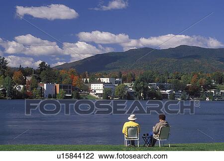 Lake Placid, NY, New York, The Adirondacks, Couple sitting on the lakefront of Mirror Lake looking at the resort town of Lake Placid in the autumn. View Large Photo Image Stock Image - Lake Placid, NY, New York, The Adirondacks, Couple sitting on the lakefront of Mirror Lake looking at the resort town of Lake Placid in the autumn.. Fotosearch