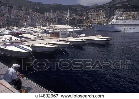 Monaco, Man fishing on a pier surrounded by boats docked in Monaco Harbor in the district of La Condamine in the Principality of Monaco along the Mediterranean Sea. View Large Photo Image Stock Photo - Monaco, Man fishing on a pier surrounded by boats docked in Monaco Harbor in the district of La Condamine in the Principality of Monaco along the Mediterranean Sea.. Fotosearch
