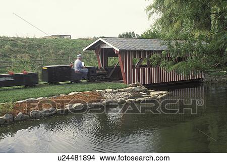 Picture -  OH, Ohio, Archbold, Small train driven by an engineer passes under a Covered Bridge at the Historic Sauder Farm and Craft Village.. Fotosearch