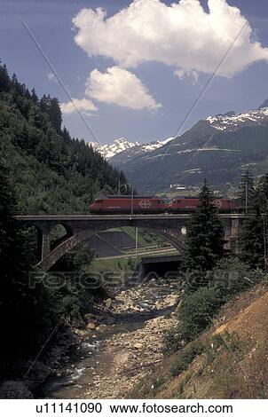 Stock Image -  train, Switzerland, Ticino, Airolo, Val Leventina, Alps, Red train crosses over a bridge through the scenic Swiss Alps.. Fotosearch