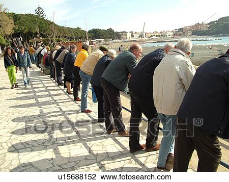 Stock Image -  bocce, Liguria, Italy, Sestri Levante, Riviera di Levante, Ligurian Riviera, Europe, Men lined up along the fence watching a game of Bocce being played on the waterfront in Sestri Levante along the Ligurian Sea.. Fotosearch