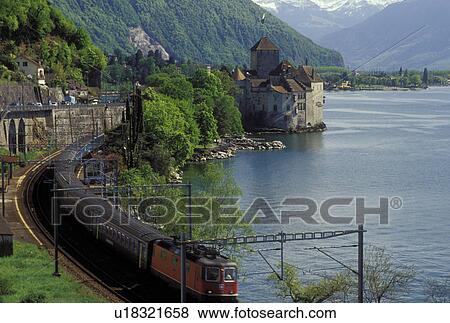 Stock Photo -  Switzerland, Montreux, Vaud, Alps, Lake Geneva, Europe, A train with a red engine winds along the lakeshore of Lac Leman with a scenic view of the Alps and the Chateau de Chillon in the spring in the Canton of Vaud. . Fotosearch