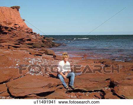 Stock Image -  Canada, Prince Edward Island, Queens County, Gulf of Saint Lawrence, Cavendish Beach, Prince Edward Island National Park, Woman sitting on red sandstone boulder along the beach. Fotosearch