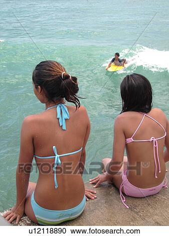 Stock Image -  Waikiki, Honolulu, Oahu, HI, Hawaii, Queen's Surf Beach, boogie boarding, 2 girls in bikini bathing suits watching. Fotosearch