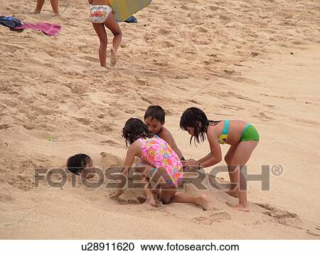 Stock Image -  Waikiki, Honolulu, Oahu, HI, Hawaii, Queen's Surf Beach, young children burying a young boy in the sand . Fotosearch