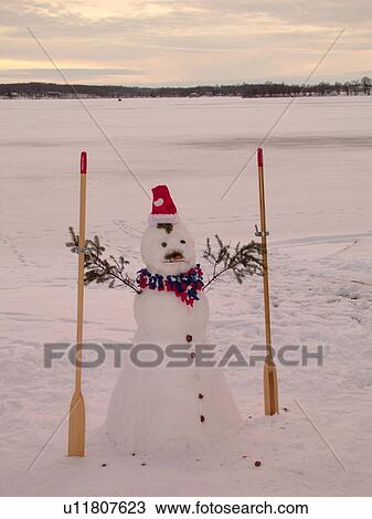Stock Image -  Alexandria, MN, Minnesota, Maple Lake, snowman wearing a red Santa hat holding oars, snow, winter. Fotosearch