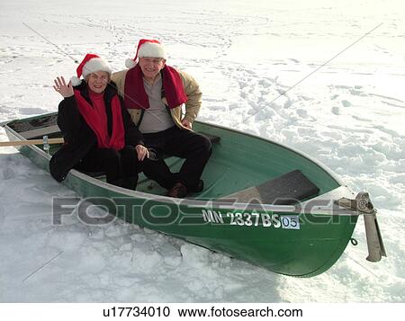 Stock Image -  Alexandria, MN, Minnesota, Maple Lake, Senior couple (husband, wife) wearing red Santa hats sitting in a green rowboat in the snow, humor, frozen lake, winter. Fotosearch