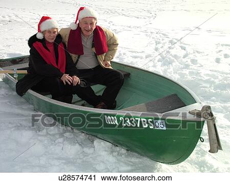 Stock Image -  Alexandria, MN, Minnesota, Maple Lake, Senior couple (husband, wife) wearing red Santa hats sitting in a green rowboat in the snow, humor, frozen lake, winter. Fotosearch