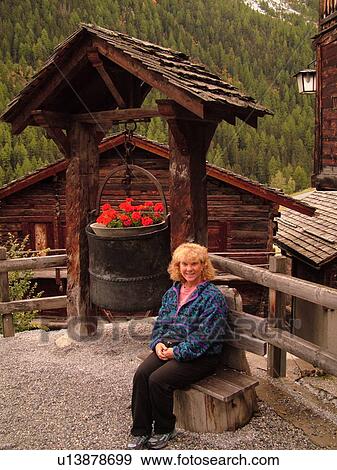 Stock Photo -  Switzerland, Europe, valais, wallis, Val d'Anniviers, Grimentz, woman sitting by a large iron kettle decorated with flowers, wishing well. Fotosearch
