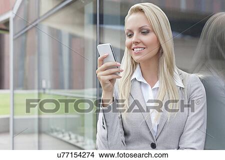Beautiful young businesswoman text messaging on mobile phone while leaning on glass wall View Large Photo Image Picture - Beautiful young businesswoman text messaging on mobile phone while leaning on glass wall. Fotosearch