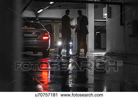 Stock Image - Full length of businessmen giving and receiving envelop in parking lot. Fotosearch