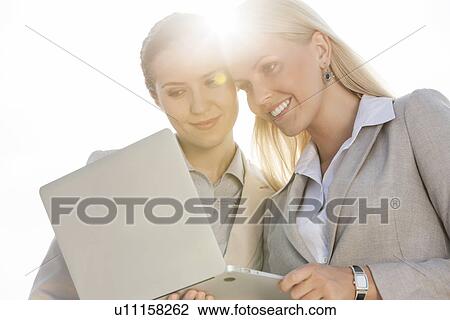 Stock Image - Happy young businesswomen using laptop against sky. Fotosearch