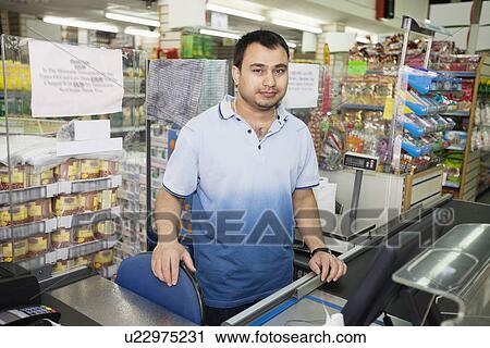 Stock Image - Portrait of male clerk standing at counter in grocery store. Fotosearch