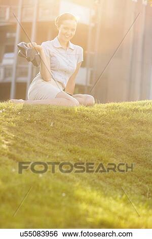 Stock Photograph - Relaxed young businesswoman holding high heels while sitting in office lawn. Fotosearch