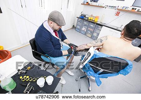 Stock Photograph - Shirtless young man getting tattooed from artist in parlor. Fotosearch
