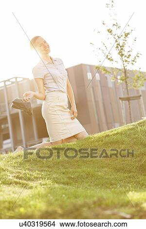 Picture - Smiling young businesswoman holding high heels while kneeling in office lawn. Fotosearch