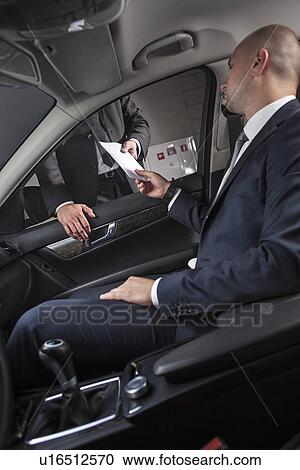 Stock Image - Young businessmen giving and receiving envelop through car window. Fotosearch