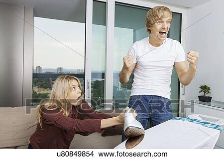Cheerful man cheering while shocked woman at the ironing board in living room View Large Photo Image Picture - Cheerful man cheering while shocked woman at the ironing board in living room. Fotosearch