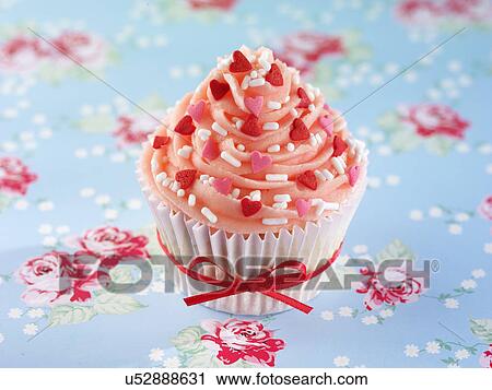 Stock Image - One Pink Buttercream Topped Cupcake With Red Heart Decorations, White Cake Case Tied With Red Ribbon, Blue, Pink And Red Floral Tabletop. Fotosearch