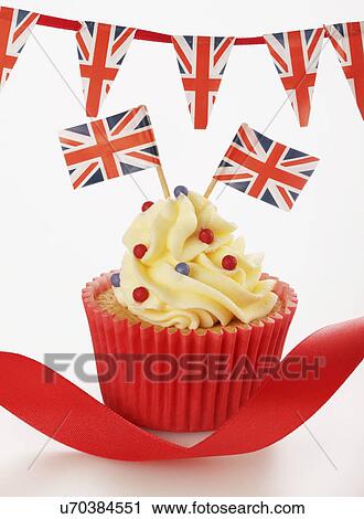 Stock Image - One Themed British Celebration Cupcake, Red Cake Case Filled With Sponge Cake, Topped With Buttercream Red And Blue Sprinkles And Small Union Jack Flags, On White With Union Jack Bunting And Red Ribbon. Fotosearch