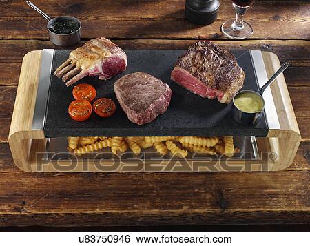 Stock Photograph - Rack of lamb rib eye and fillet steak on a hot lava stone with chips underneath. Fotosearch