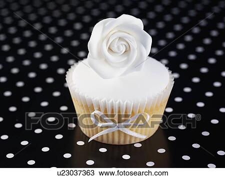 Stock Image - Single White Iced Cupcake With White Rose On Top, Black Polka Dot Tabletop. Fotosearch