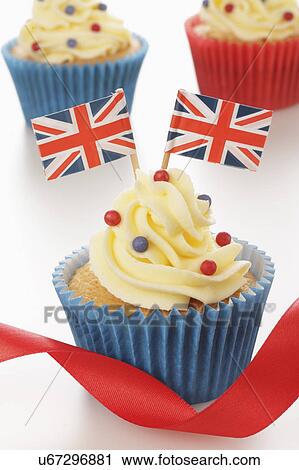 Stock Image - Themed British Celebration Cupcake, Blue Cake Case With Sponge Cupcake, Topped With Buttercream Red And Blue Sprinkles And Two Small Union Jack Flags, On White With Red Ribbon And Two Cakes In Background. Fotosearch